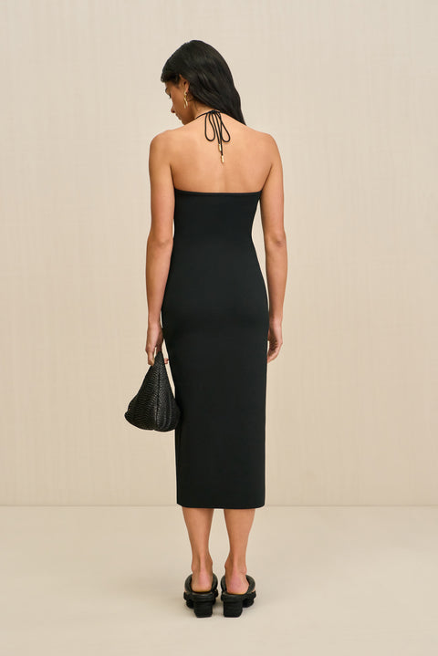 Back facing woman wearing a black midi dress that features an adjustable halter neck. 