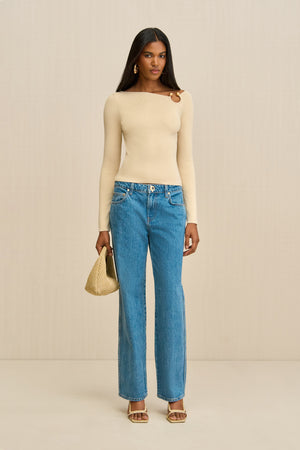 Woman wearing a cream long sleeve shirt with a ring detail at the shoulder, paired with blue jeans. 
