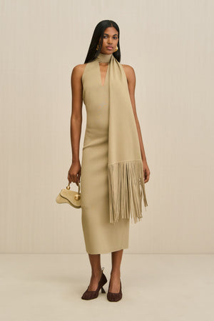 Woman wearing a tan, knit midi dress that features a V neck and a detachable fringe scarf, paired with a gold handbag and brown heels. 