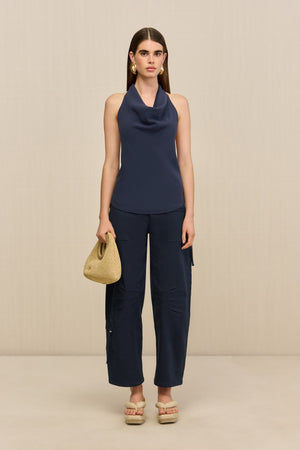 Front-facing woman wearing a navy blue sleeveless top that features a cowl halter neck, paired with blue pants, natural sandals, and a woven handbag. 