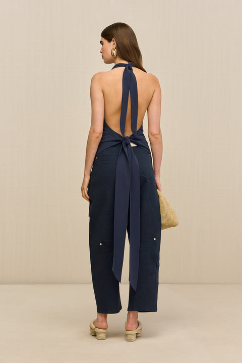 Back-facing woman wearing a navy blue sleeveless top that features an adjustable tie halter neck and a low, open back secured with another adjustable tie around the hips, paired with navy pants and tan sandals. 