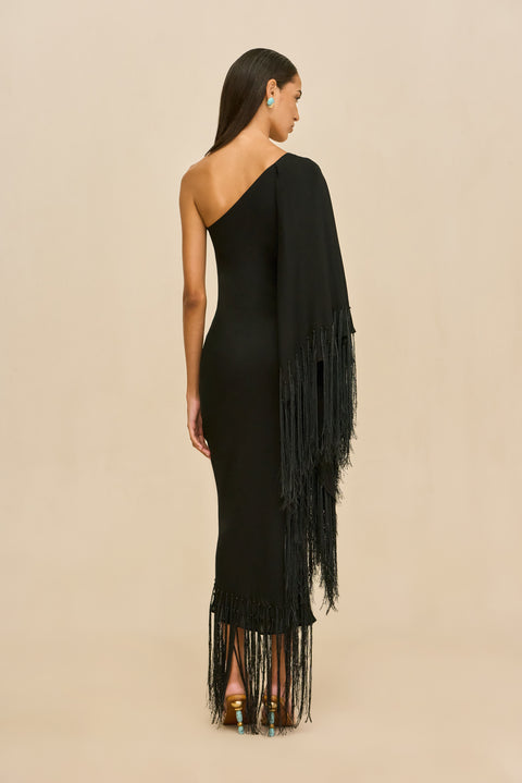 Back-facing shot of woman wearing a black one-shoulder dress with fringe details on a beige background