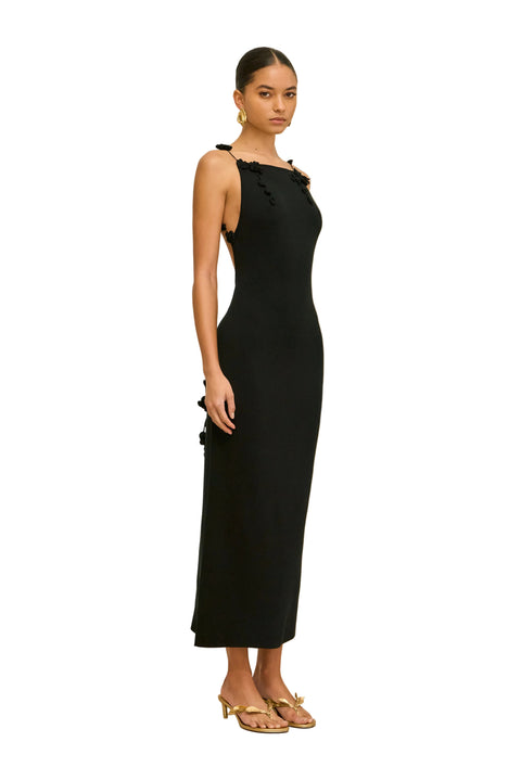 Woman wearing a black sleeveless dress with hanging floral straps on a beige background standing to the side