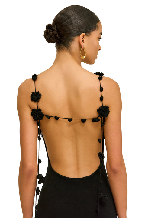 Back-facing shot of woman wearing a black sleeveless dress with hanging floral straps on a beige background