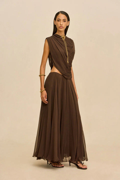 Woman wearing a brown outfit with a draped top and long skirt on a beige background standing off-center.