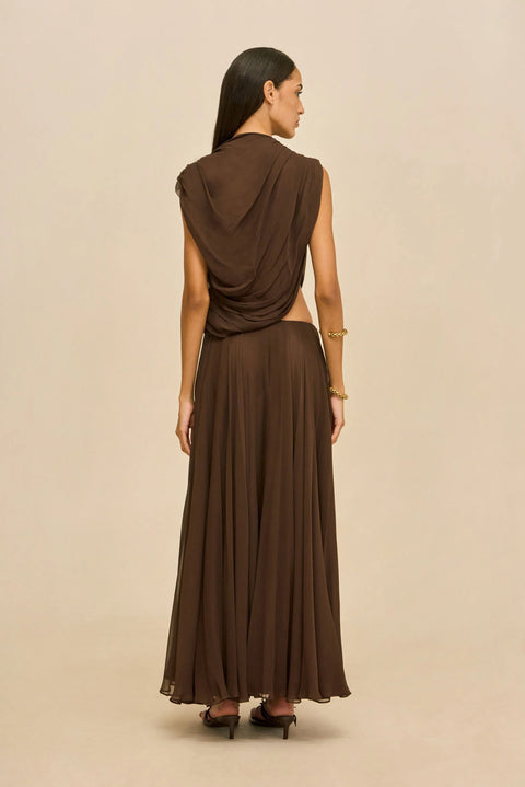 Back-facing shot of woman wearing a brown outfit with a draped top and long skirt on a beige background