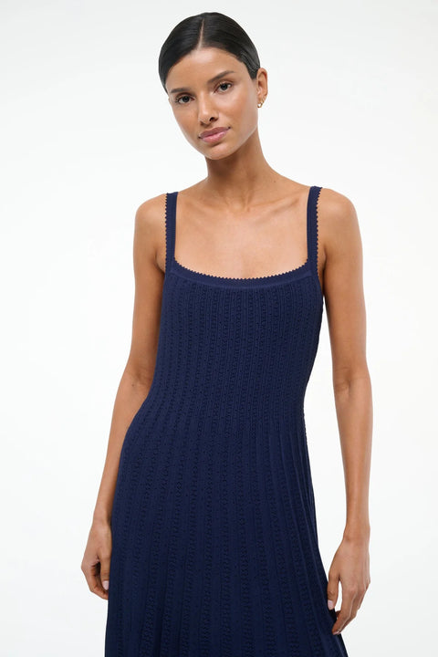 Close up of a woman wearing a sleeveless, navy knit midi dress that features a square neck, ribbed, fitted bodice, and pleated skirt. 