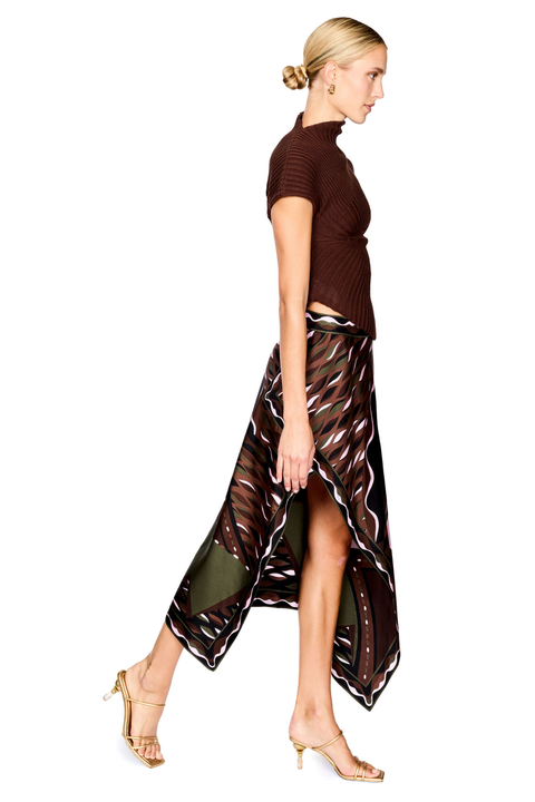 A model stands against a white studio background wearing a dark brown ribbed short-sleeve mock-neck top and a high-low wrap skirt with a black, brown, beige, and pink abstract print. She is holding a small blush pink handbag and wearing gold high-heeled sandals.