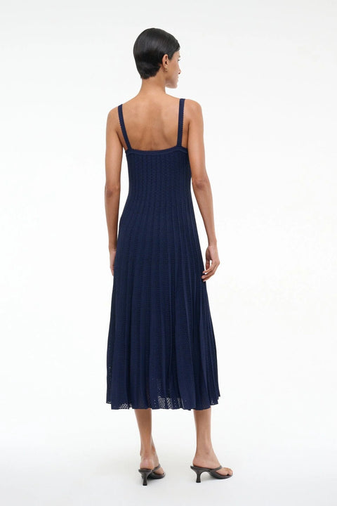 Back view of a woman wearing a sleeveless, navy midi dress that features a square back, fitted bodice, and pleated skirt, paired with black heels. 