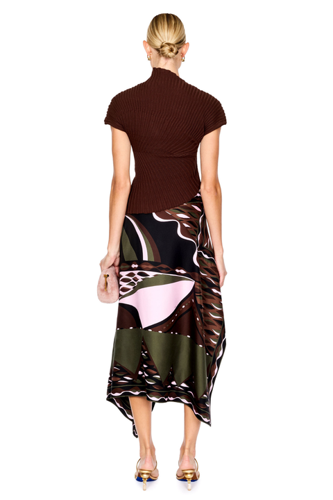 A model stands against a white studio background wearing a dark brown ribbed short-sleeve mock-neck top and a high-low wrap skirt with a black, brown, beige, and pink abstract print. She is holding a small blush pink handbag and wearing gold high-heeled sandals.