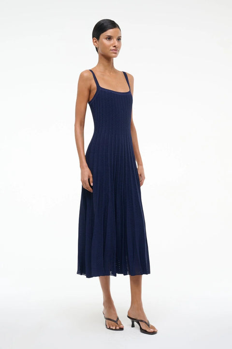 Side view of a woman wearing a sleeveless, navy knit midi dress that features a square neck, fitted bodice, and peated skirt, paired with black heels. 