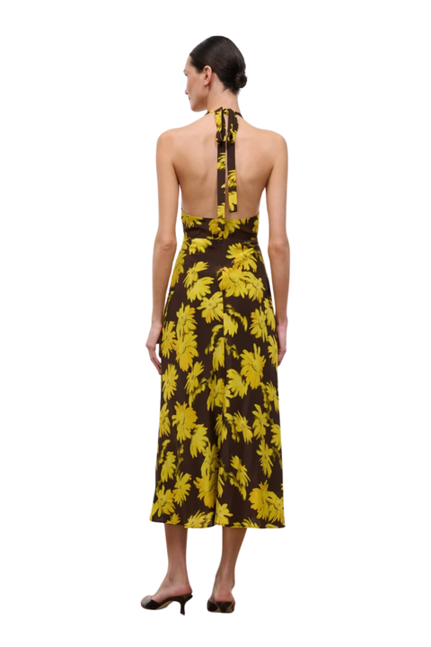 A woman shown from the back wearing a halter-neck midi dress with a yellow tropical leaf print on a dark brown background, paired with black heels, against a plain white studio background.