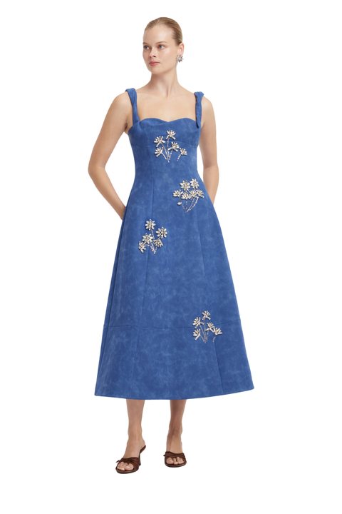 A woman modeling a sleeveless blue midi dress with white floral embroidery, photographed against a plain white background.