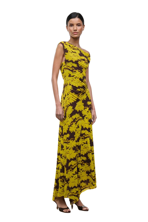 A woman modeling a sleeveless, one-shoulder maxi dress with a bold yellow and dark brown abstract floral pattern, set against a plain white background.
