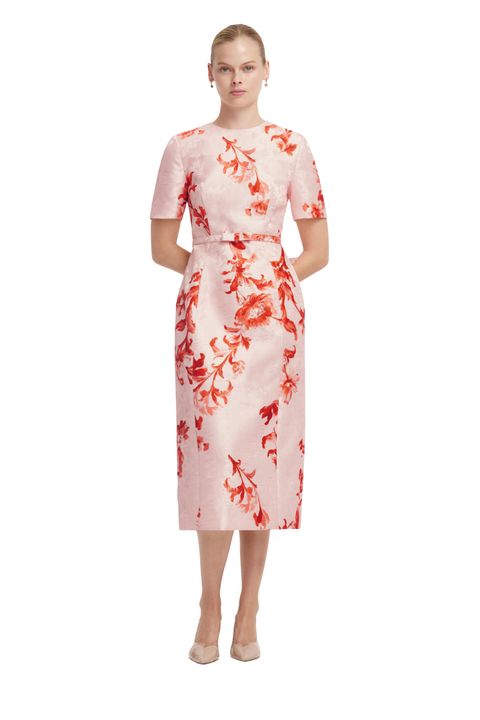 A woman modeling a short-sleeved midi dress with a pink base and a red-orange floral pattern, standing against a plain white background.