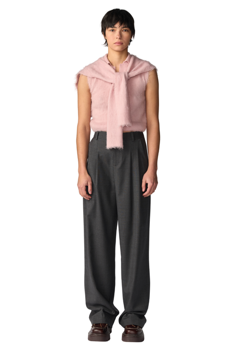 A model stands against a white background wearing a light pink sleeveless fuzzy top with a draped scarf detail, paired with dark gray pleated trousers and brown shoes.