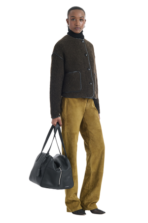 Person wearing a brown jacket and mustard pants holding a black bag on a white background