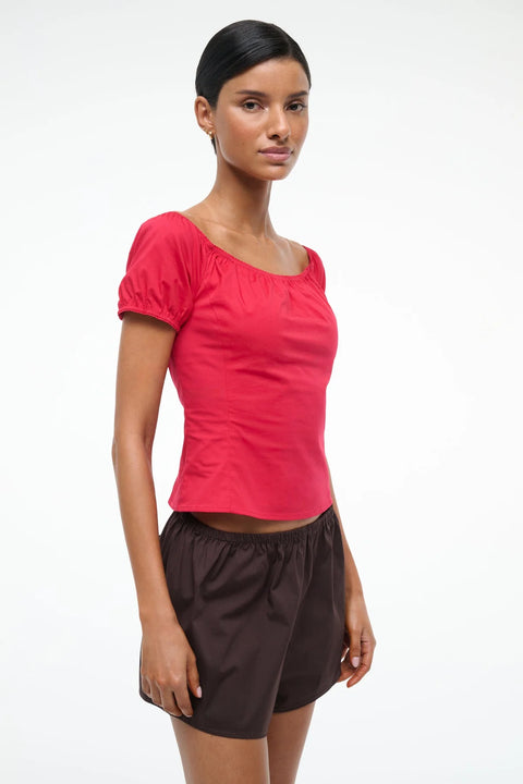 Woman wearing a bright red top with puff short sleeves, which can be worn on or off the shoulder, paired with brown shorts.