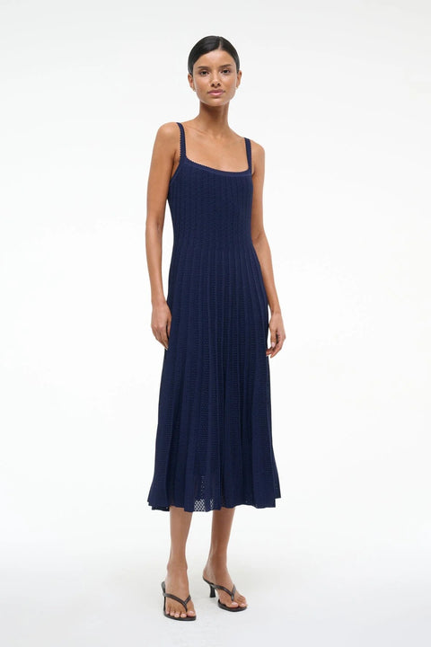 Woman wearing a sleeveless, navy knit midi dress that features a square neck, ribbed, fitted bodice, and pleated skirt, paired with black heels. 