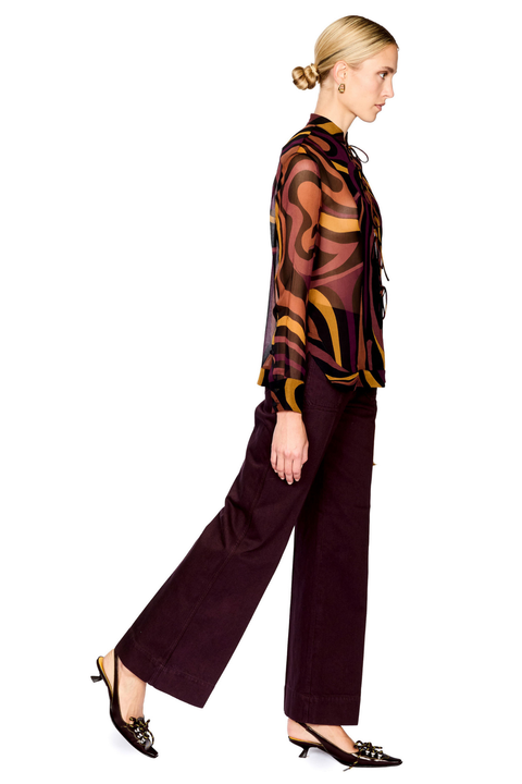 A woman modeling a sheer long-sleeve button-up blouse with an abstract wavy pattern in brown, gold, and black, paired with dark wide-leg trousers and brown slingback heels, shown against a plain white background.