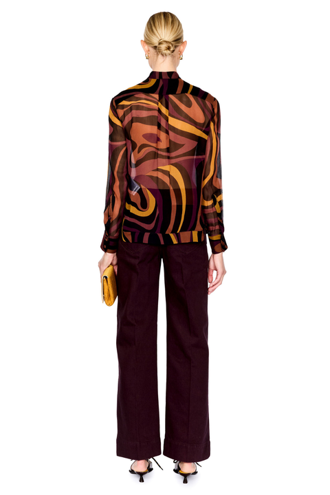 A model shown from the back wearing a sheer long-sleeve top with an abstract wavy pattern in brown, orange, and purple, paired with dark plum wide-leg trousers and holding a small yellow clutch against a white background.