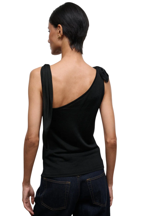 A woman shown from the back wearing a black asymmetrical one-shoulder top with a draped strap and a bow detail on the other shoulder, paired with dark blue jeans against a white background.