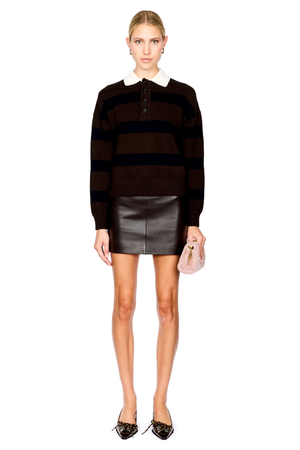 A model stands against a white studio background wearing a dark brown and navy horizontally striped knit polo sweater with a white collar, a black leather mini skirt, and brown pointed flats, while holding a small pale pink clutch.