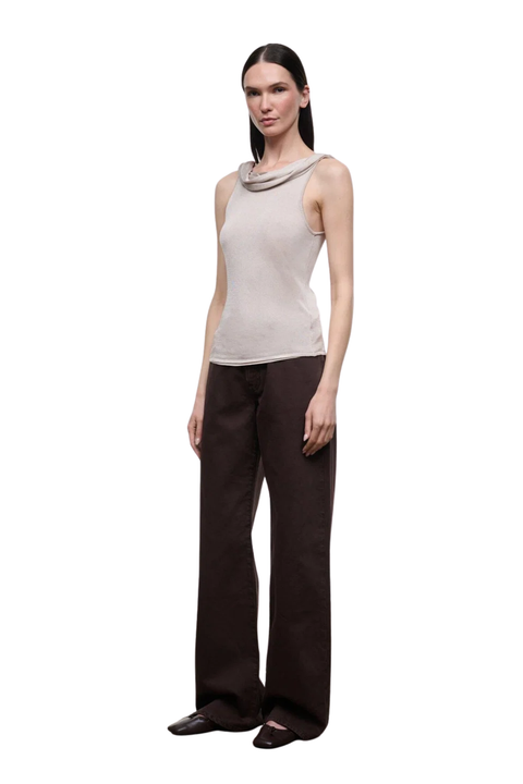A woman modeling a sleeveless champagne colored top with a draped cowl neckline and a side tie detail, paired with dark brown pants.