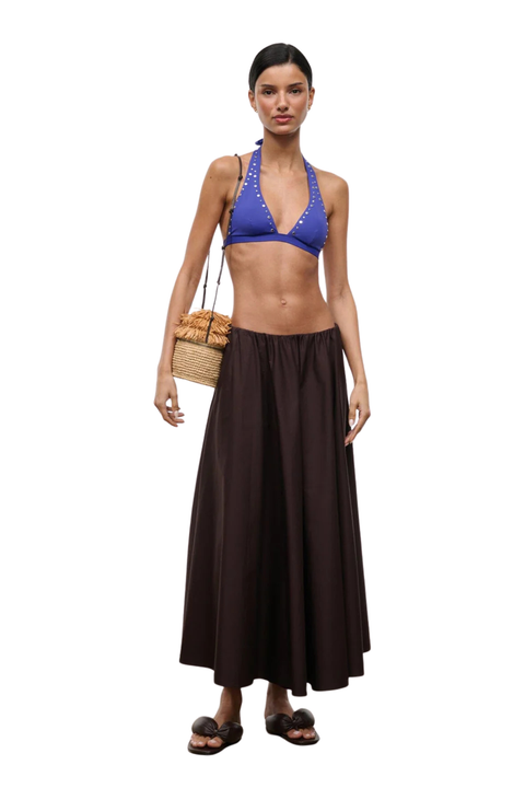 A model stands against a white background wearing a two-piece swim set consisting of a royal blue halter bikini top with white trim and a dark brown maxi skirt. She is also carrying a woven straw bag and wearing dark brown slide sandals.