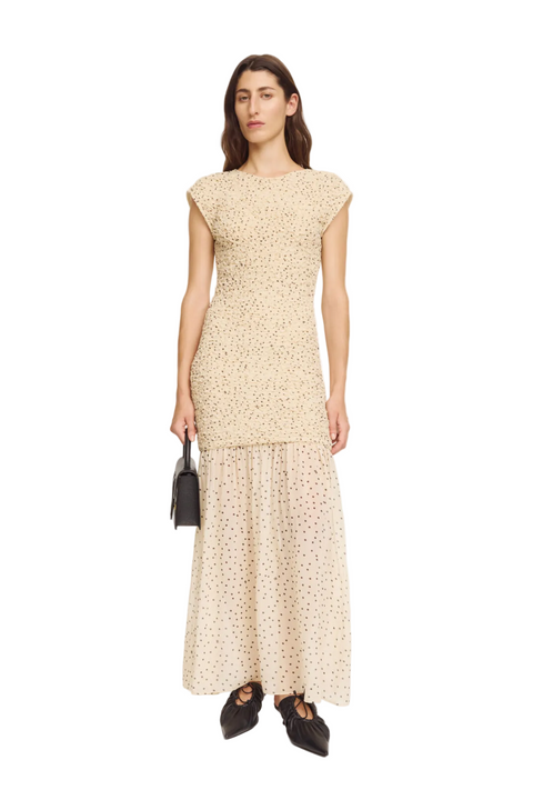 Woman wearing a beige patterned dress holding a black handbag on a white background.
