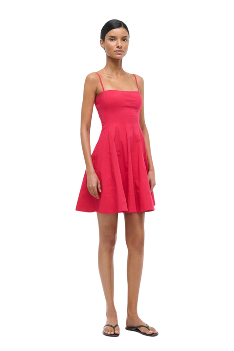 A woman modeling a bright cherry red sleeveless mini dress with thin straps and a flared skirt, standing against a plain white background.