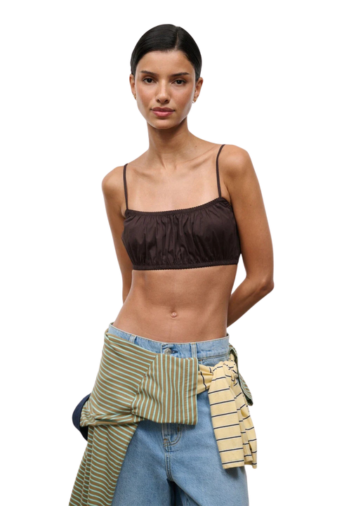 A model wearing a dark brown ruched spaghetti-strap crop top, styled with light-wash blue jeans and a striped green and yellow shirt tied around her waist, against a plain white background.