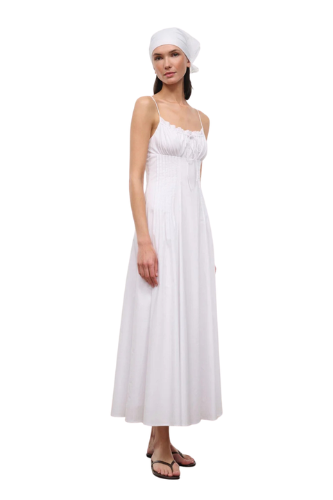 A woman modeling a long white maxi dress with thin straps, a gathered bodice, and a flowing skirt, paired with a white headscarf and dark sandals, against a plain white background.
