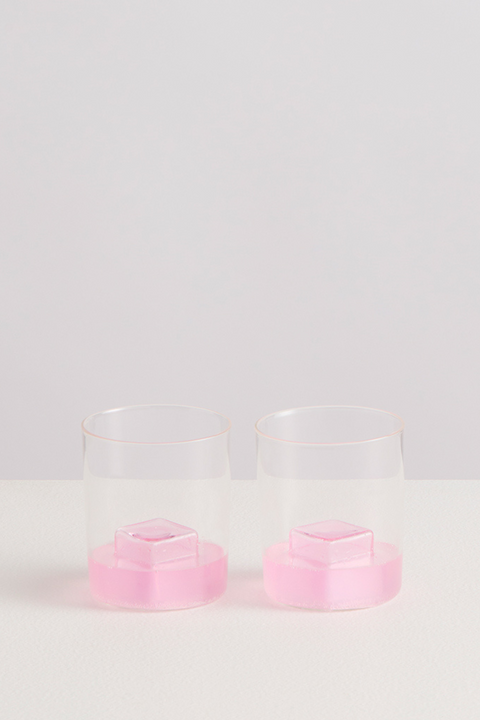 Clear glasses with a little pink liquid with pink glass ice cubes inside them on a light gray background