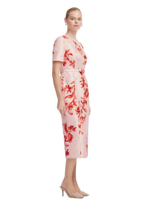 A woman modeling a short-sleeve midi dress with a red floral pattern on a light pink textured fabric, paired with beige pointed-toe heels, against a plain white background.