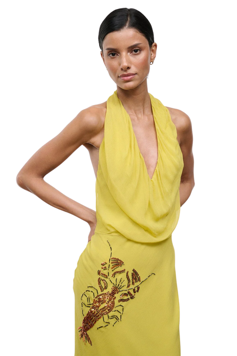 A woman modeling a bright yellow halter-neck dress with a draped cowl neckline and a red lobster sequin embellishment on the lower left side, set against a plain white background.