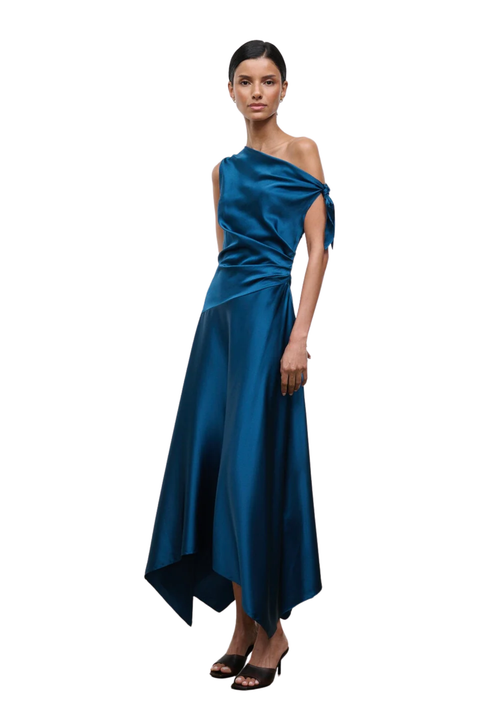 A model wearing a dark teal blue satin one-shoulder midi dress with a draped, gathered skirt, paired with black heeled sandals, set against a plain white background.