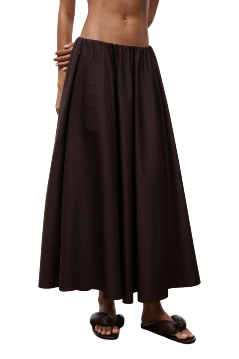 A dark brown, flowy maxi skirt worn by a model, paired with dark brown slide sandals, shown against a plain white background.