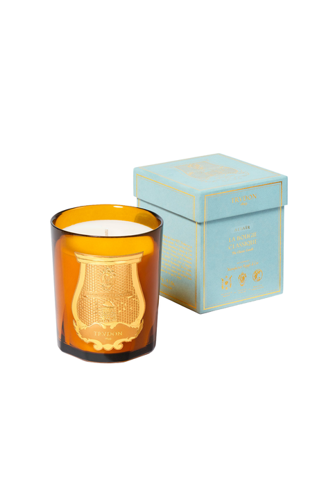 An amber murano glass jar candle with a gold crest emblem on the front, containing white wax and a light blue gift box, set against a plain white background.