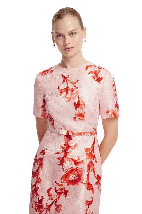 A woman modeling a light pink short-sleeve dress with a red floral pattern and a matching waist tie, set against a plain white background.