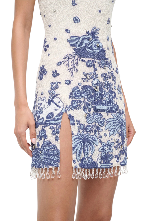 A close-up of a model wearing a sleeveless bodycon mini dress with a blue and white toile-style pattern and a fringe tassel hem.