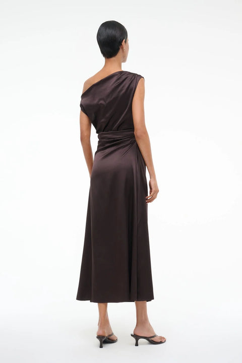 Back view of a woman wearing an asymmetrical brown silk midi dress with an off the shoulder design and draped ruching throughout, paired with brown heels 