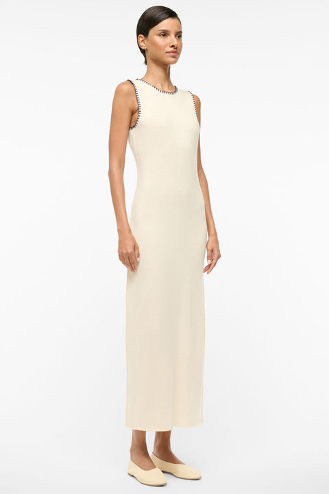 Woman wearing a sleeveless ivory maxi dress that features navy whipstitch detailing around the arm holes and neck holes. 