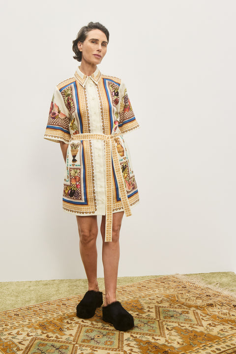 Woman wearing a cream and multicolor, printed, mini shirt dress that features a point collar and a self-tie belt, paired with black mules. 