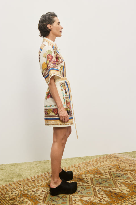 Side-facing woman wearing a cream and multicolor, printed, mini shirt dress that features a point collar and a self-tie belt, paired with black mules. 