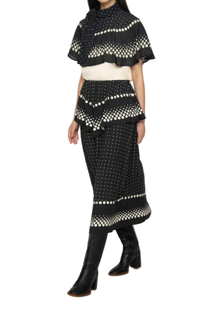 A model wearing a black and white dotted tiered top with a high neck, short flutter sleeves, a cream waistband, and black knee-high boots, set against a plain light gray background.