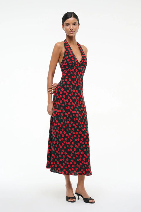 Side view of a woman wearing a black and red tomato print midi dress that features a halter neck and deep V, paired with black heels. 