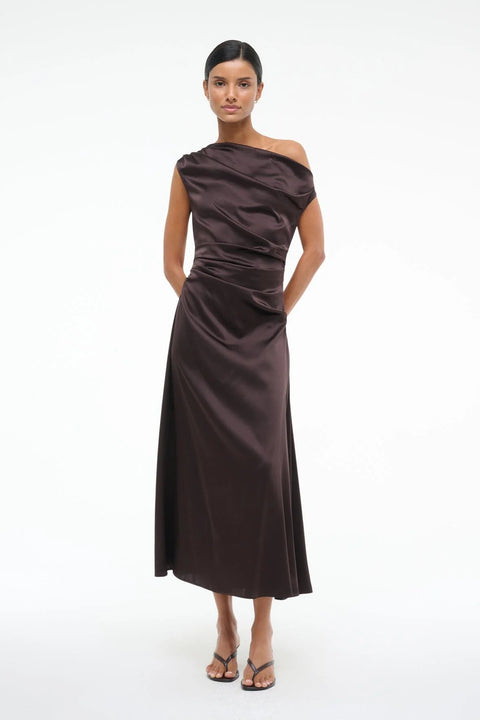 Woman wearing an asymmetrical, brown silk midi dress that features an off the shoulder design with draped ruching throughout, paired with brown sandals. 