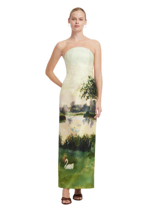 A woman modeling a strapless maxi dress with a landscape print featuring green reeds, a tree, water, and a white swan, set against a plain white background.