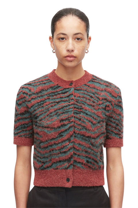 A model wearing a short-sleeve button-up cardigan with a textured zebra-like pattern in rust red and dark teal, paired with black trousers, shot against a plain white background.